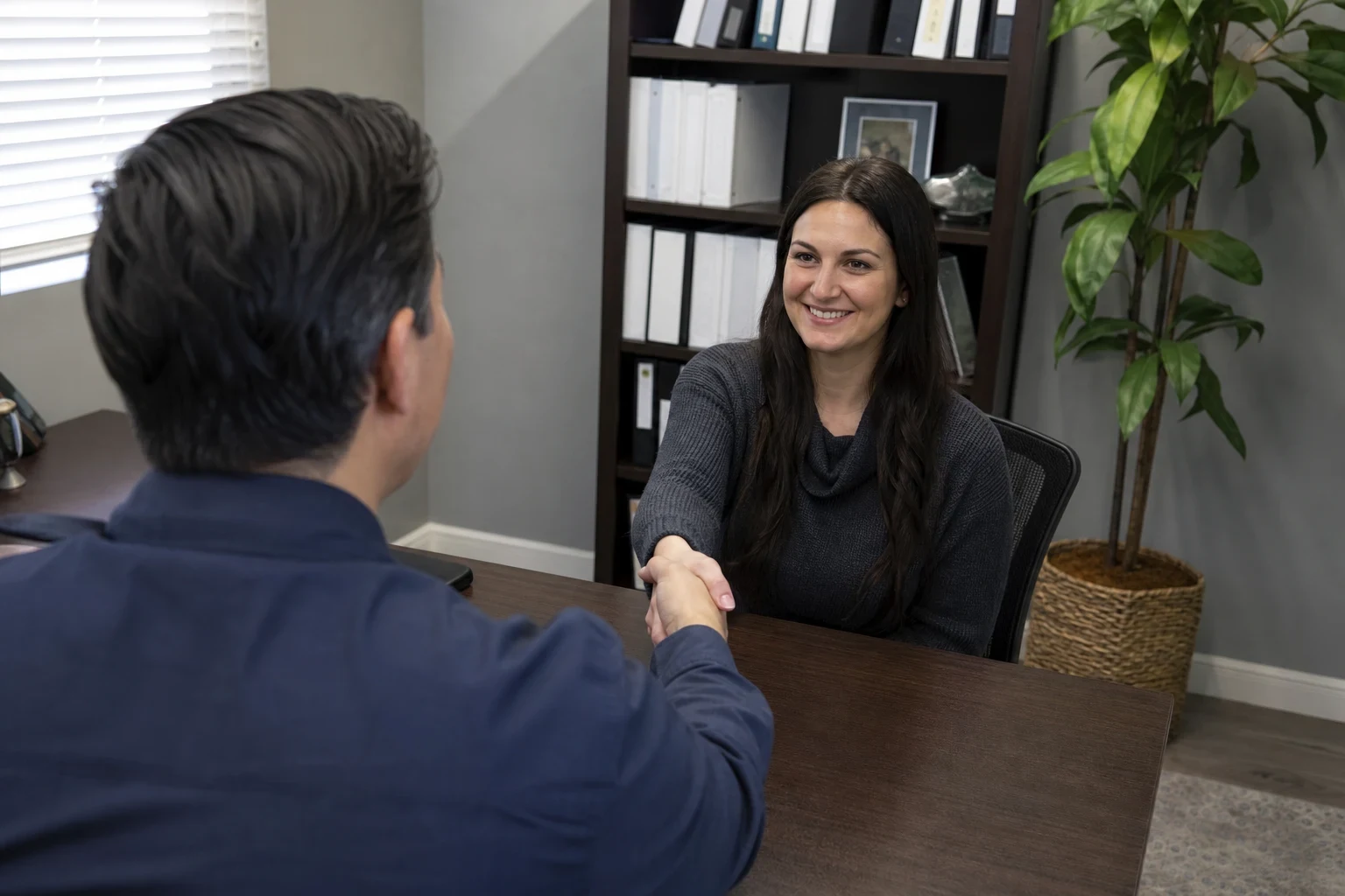 Man in a navy shirt shakes hands across a wooden desk with a smiling woman in a gray sweater, with a bookshelf of binders and a potted plant behind her