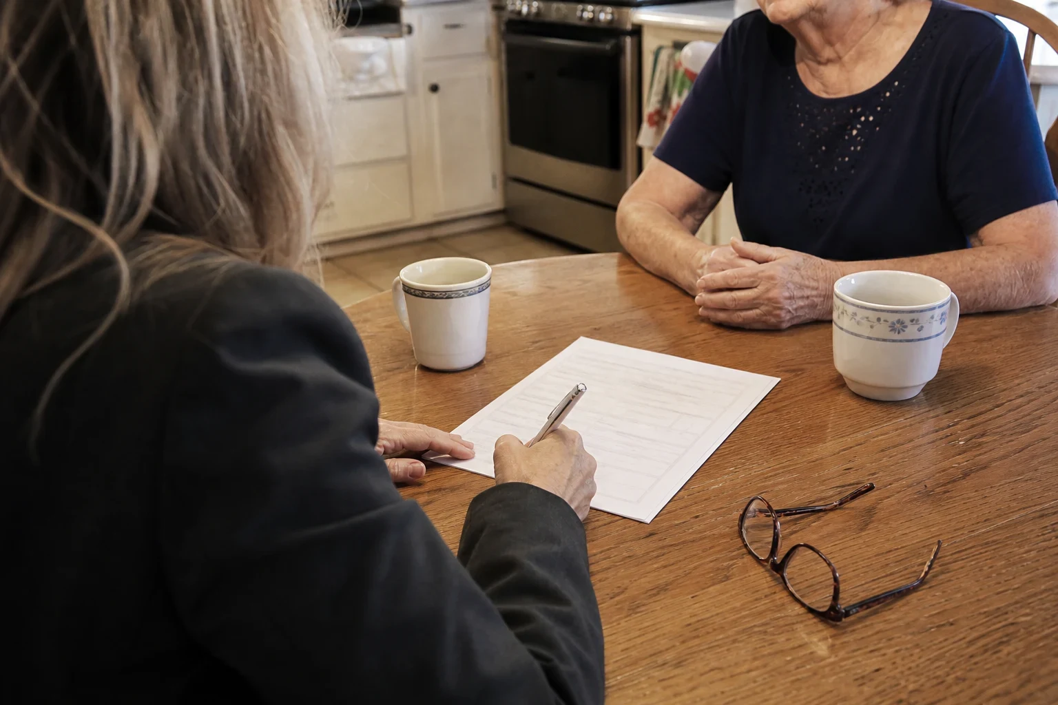 Agent filling out a form at a wooden kitchen table across from an older woman, with two coffee mugs and a pair of reading glasses on the table