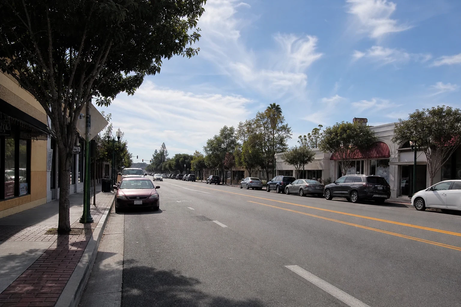 Wide view of a sunny downtown street lined with low storefronts, red awnings, street trees and parked cars along both curbs under a lightly clouded blue sky.